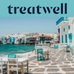 El paseo marítimo de la Pequeña Venecia en Mykonos: casas encaladas con balcones de madera de colores al borde del agua, el mar Egeo turquesa al fondo y, en primer plano, mesas de café blancas y verde azulado dispuestas sobre una terraza adoquinada.