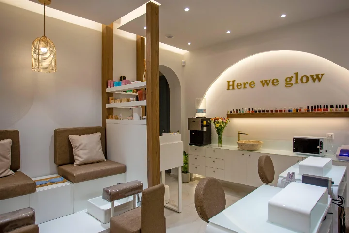 Un salón de uñas cálido y de luz suave, con una lámpara colgante de ratán trenzado, bancos de pedicura marrones acolchados, mesas de manicura blancas, una estantería de esmaltes de colores y un cartel dorado «Here we glow» sobre un arco retroiluminado.