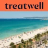 Vista aérea de la Playa de Palma en Mallorca, con una larga curva de arena dorada, hileras de sombrillas, agua turquesa en calma y un paseo bordeado de palmeras junto a la ciudad, con colinas a lo lejos bajo un cielo azul despejado.