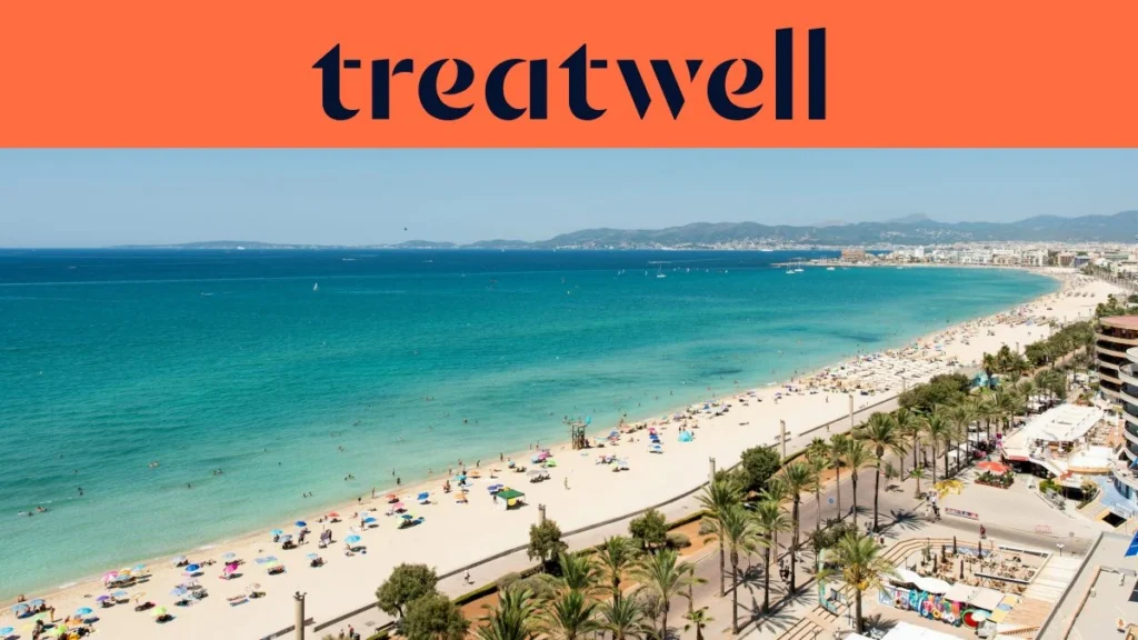 Vista aérea de la Playa de Palma en Mallorca, con una larga curva de arena dorada, hileras de sombrillas, agua turquesa en calma y un paseo bordeado de palmeras junto a la ciudad, con colinas a lo lejos bajo un cielo azul despejado.