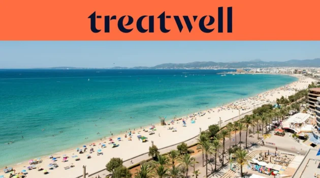 Vista aérea de la Playa de Palma en Mallorca, con una larga curva de arena dorada, hileras de sombrillas, agua turquesa en calma y un paseo bordeado de palmeras junto a la ciudad, con colinas a lo lejos bajo un cielo azul despejado.
