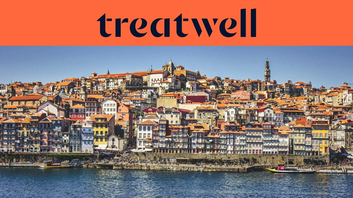 El paseo fluvial de la Ribeira en Oporto, con hileras de casas de colores y tejados de teja que ascienden por la ladera desde el río Duero, y la torre de los Clérigos en el horizonte bajo un cielo azul despejado.