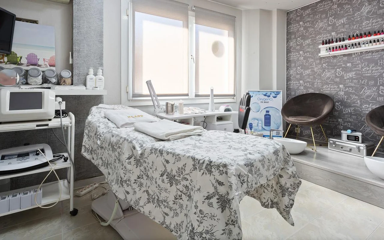 Una luminosa sala de tratamientos de belleza con una camilla vestida con ropa de cama de flores grises y toallas blancas dobladas, aparatos de estética en carritos a la izquierda, una ventana con la persiana medio bajada y una zona elevada con una butaca de terciopelo marrón y barreños de pedicura contra una pared decorativa con estampado de letras.
