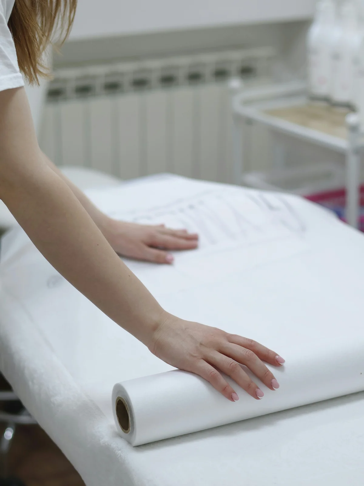 Primer plano de los brazos de una esteticista que despliega una sábana de papel limpia sobre una camilla para preparar un tratamiento