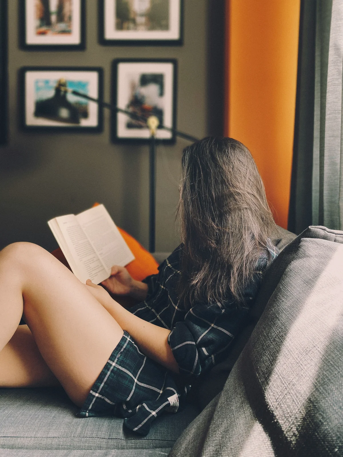 Vista lateral de una mujer sentada en un sofá leyendo un libro, con el pelo largo cubriéndole la cara; lleva unos shorts holgados de algodón y una camisa de cuadros verde oscuro