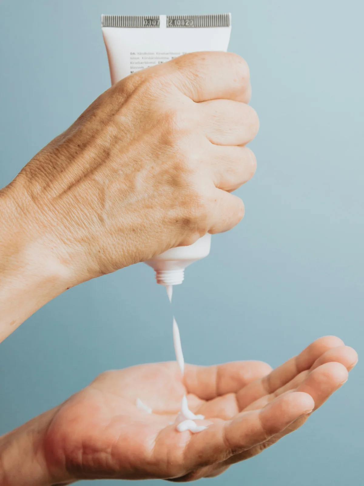 Primer plano de una mano que sujeta en vertical un tubo de crema hidratante blanca y echa un poco en la otra mano, debajo