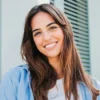 Una mujer de pelo largo y oscuro con las cejas bien definidas sonríe al aire libre frente a un edificio con contraventanas verdes