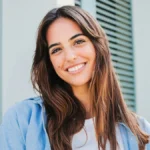 Una mujer de pelo largo y oscuro con las cejas bien definidas sonríe al aire libre frente a un edificio con contraventanas verdes
