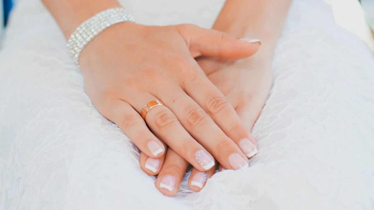 Las manos de una mujer reposan sobre su regazo; lleva un vestido de novia de encaje y una cuidada manicura francesa en una combinación clásica de nude y blanco sobre uñas cortas cuadradas-ovaladas, con un delicado adorno en los anulares