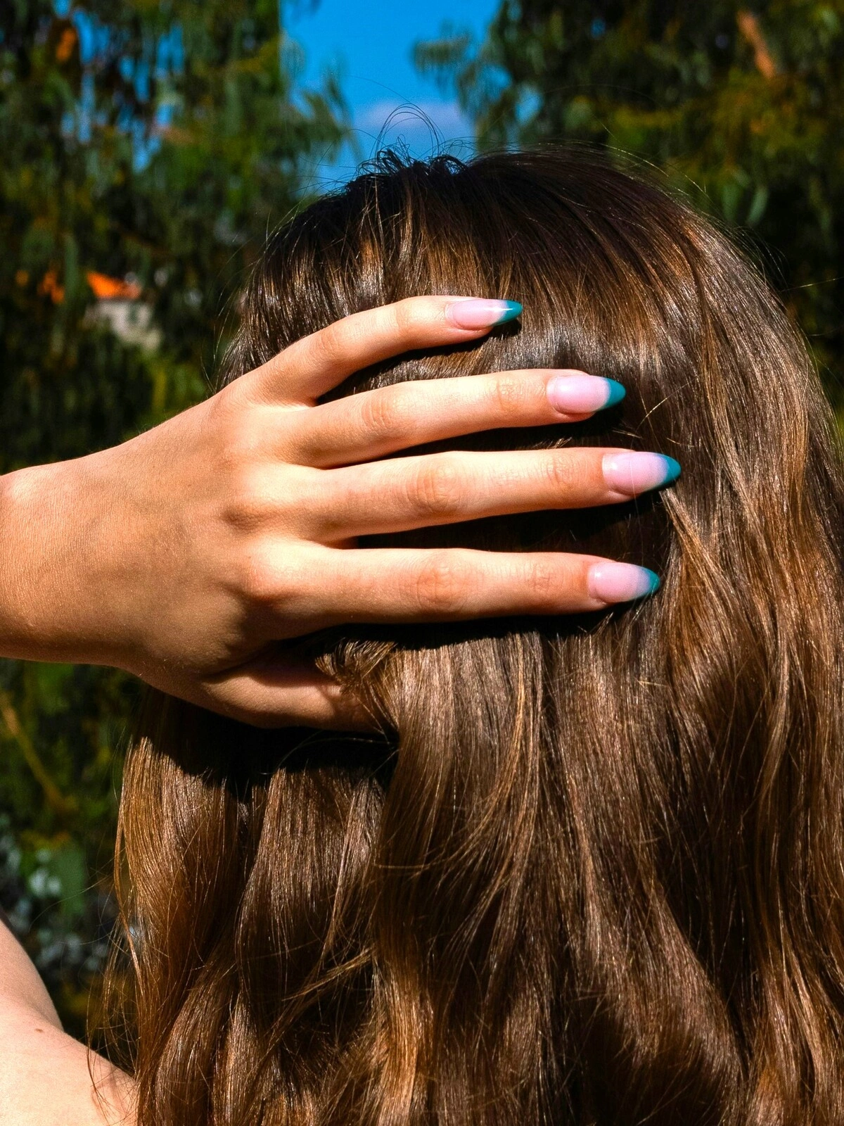 La mano de una mujer apoyada en la nuca muestra una manicura francesa de base rosa claro y puntas en verde azulado sobre uñas de almendra