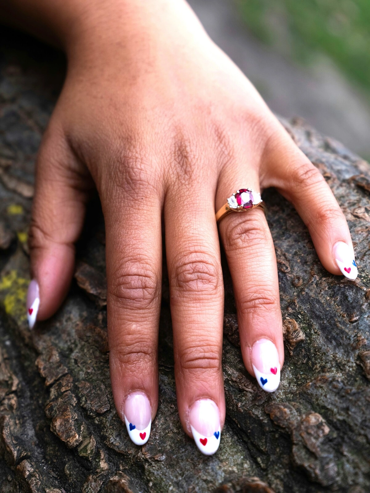 La mano de una mujer apoyada sobre una rama muestra una manicura francesa sobre uñas ovaladas, decoradas con pequeños corazones rojos y azules