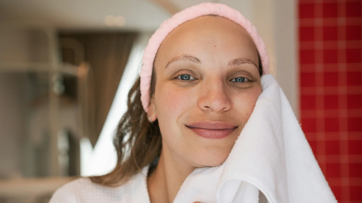 Una mujer con la piel limpia y luminosa sonríe a la cámara con una diadema rosa mientras se seca el rostro recién limpio con una toalla blanca