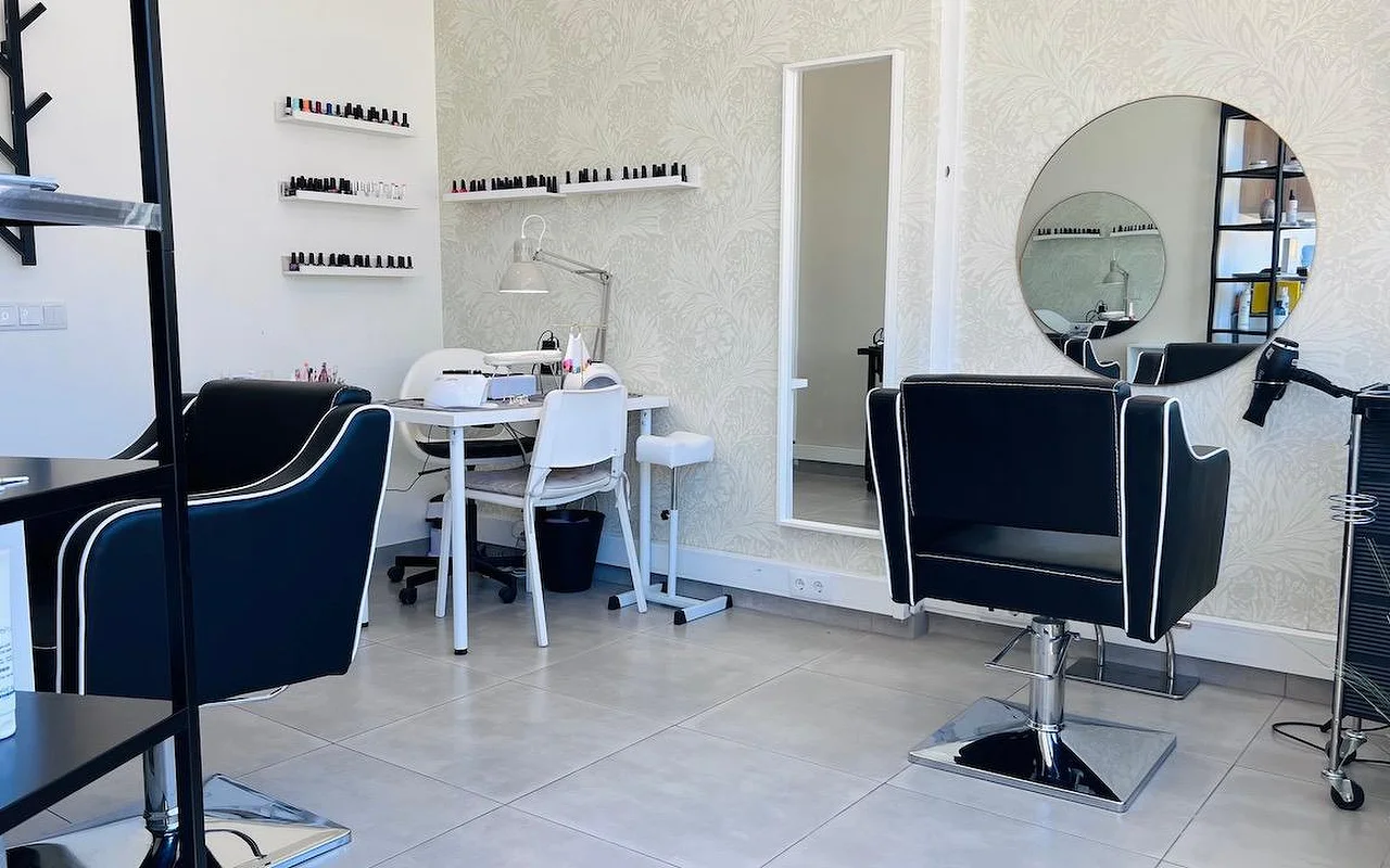 Un compacto salón de pelo y uñas con sillones de peinado negros ribeteados en blanco, un espejo redondo y un espejo alto sobre un papel pintado gris de flores, y estanterías de pared alineadas con esmaltes.