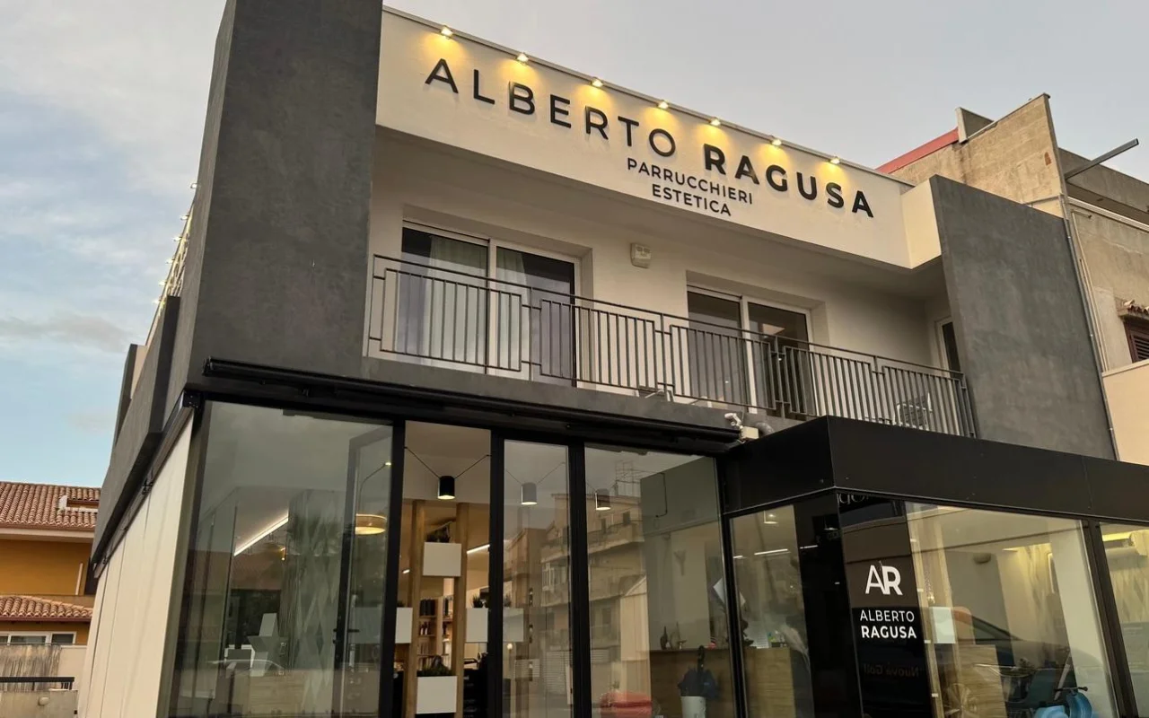 El exterior de Alberto Ragusa al anochecer, un salón de dos plantas con un gran letrero iluminado que dice «Alberto Ragusa Parrucchieri Estetica» sobre ventanales del suelo al techo, con una luz cálida que sale del interior.