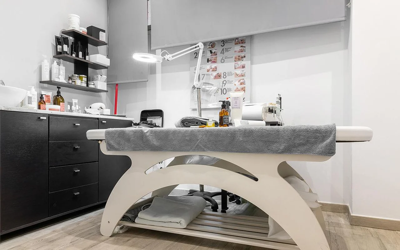 Una cabina de estética compacta en Atelier Beauty and the Lab, con una camilla color crema, una lámpara-lupa regulable, estantes oscuros en la pared con productos para la piel y, en la pared, un esquema numerado de los pasos del tratamiento.