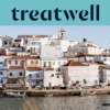 Un pueblo pesquero del Algarve encalado en blanco, con casas de tejados de terracota que ascienden por una ladera sobre una ría en calma, pequeñas barcas de pesca amarradas junto al muelle, palmeras y el campanario de una iglesia bajo un cielo azul despejado.