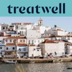Un pueblo pesquero del Algarve encalado en blanco, con casas de tejados de terracota que ascienden por una ladera sobre una ría en calma, pequeñas barcas de pesca amarradas junto al muelle, palmeras y el campanario de una iglesia bajo un cielo azul despejado.