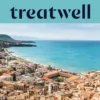 Vista panorámica de un pueblo costero siciliano, con antiguos edificios de tejados de terracota, una media luna de playa de arena, un mar turquesa cristalino y un promontorio rocoso que se alza a un lado.