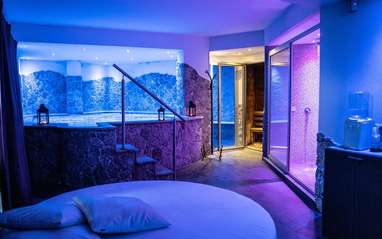 Una suite de spa privada en Bellezza e Benessere, en Taormina, iluminada de azul y morado, con una bañera de hidromasaje empotrada en un revestimiento de piedra, una sauna de madera y, al fondo, un baño turco de mosaico, y una cama redonda en primer plano.