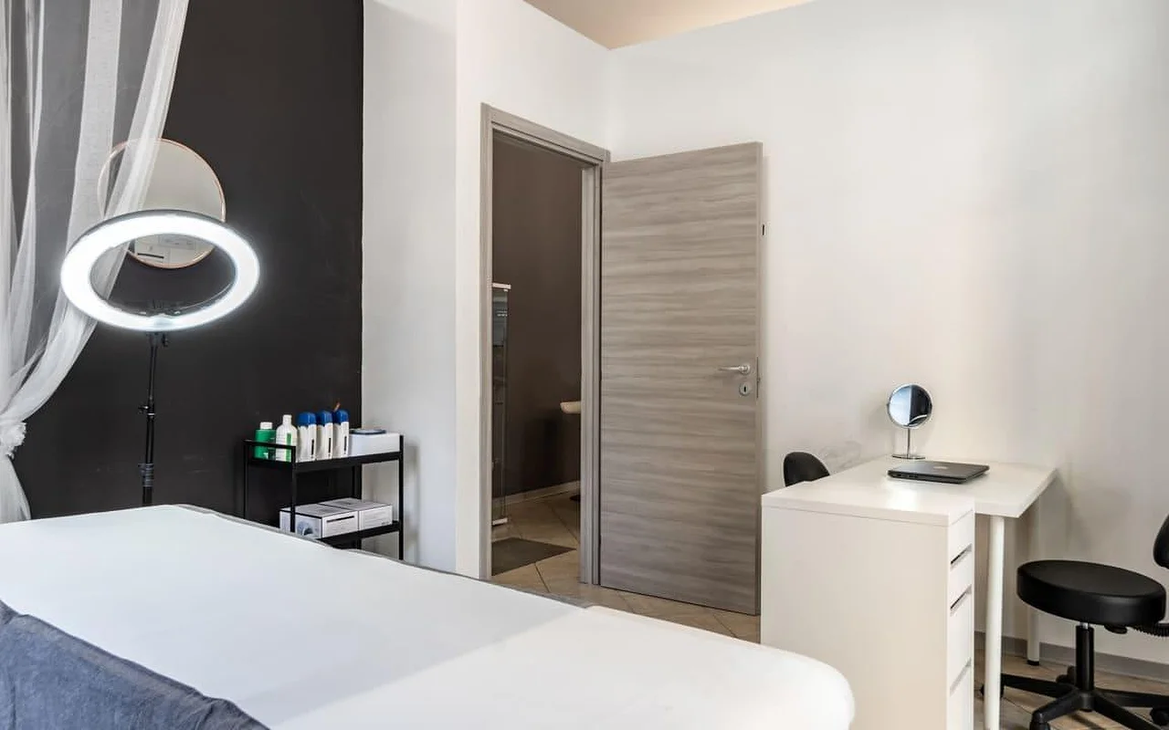 Una cabina de tratamiento en Coquette Wellness and Beauty, con una camilla blanca, un aro de luz encendido sobre un pie contra una pared negra decorativa, visillos en la ventana y un pequeño escritorio blanco con un espejo de tocador junto a una puerta que da a un baño privado.