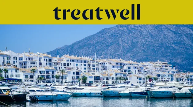 El puerto deportivo de Puerto Banús, en Marbella, rodeado de edificios blancos y yates amarrados con las montañas al fondo, bajo una banderola amarilla de Treatwell.