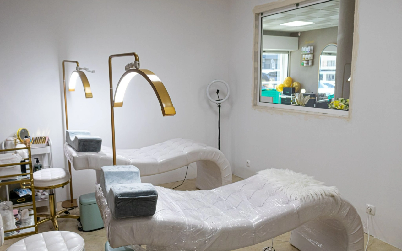 Una luminosa sala de tratamientos beauty con dos camillas blancas acolchadas, altas lámparas de arco doradas y un aro de luz, con una ventana interior que da a la recepción.