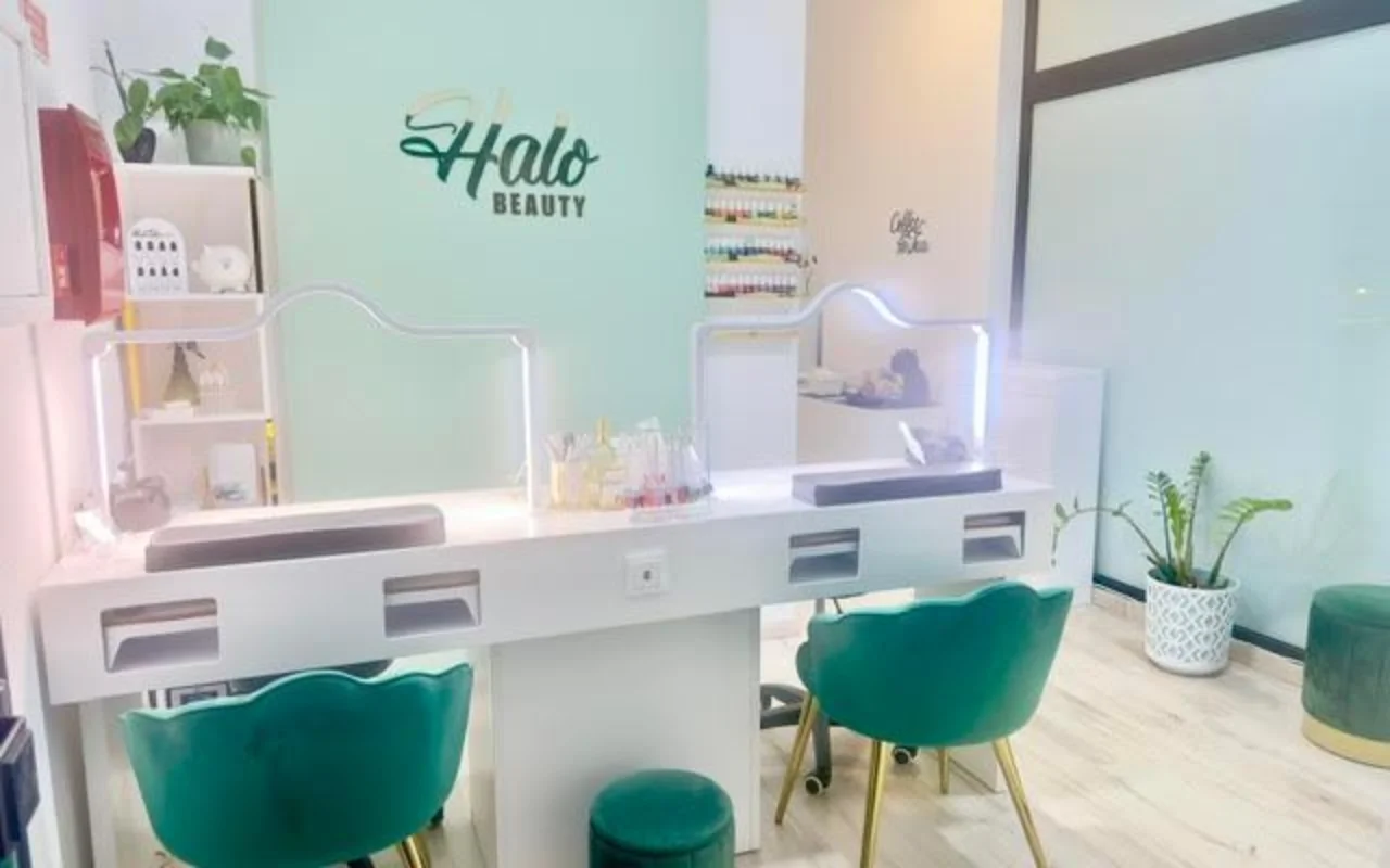 Un salón de uñas con una pared decorativa verde menta que luce el logo de Halo Beauty, un doble mostrador de manicura blanco con separadores de metacrilato iluminados, dos sillones tipo tonel de terciopelo verde esmeralda, un expositor de esmaltes en la pared y plantas en macetas.