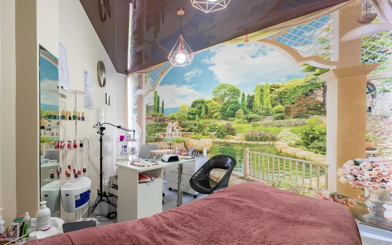Una sala de tratamientos beauty con una camilla cubierta de rosa y un puesto de uñas blanco, ante un gran mural de pared de jardín y lago, con estanterías de vidrio surtidas de productos al lado.