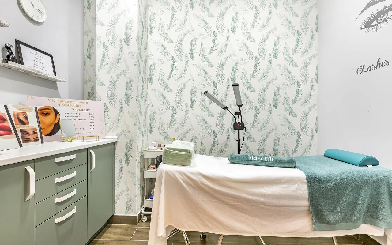 Una sala de tratamiento en Masami Belleza Natural en Fuengirola, con una camilla, papel pintado de plumas, una lámpara lupa y un cartel de precios de micropigmentación.