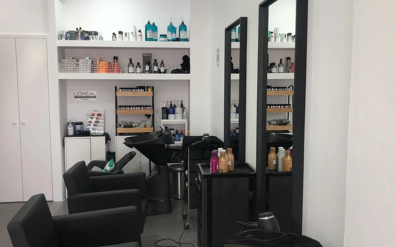 Una peluquería con sillones y lavacabezas negros, dos espejos de pared altos y estrechos, y estanterías surtidas de productos profesionales de color y cuidado capilar.