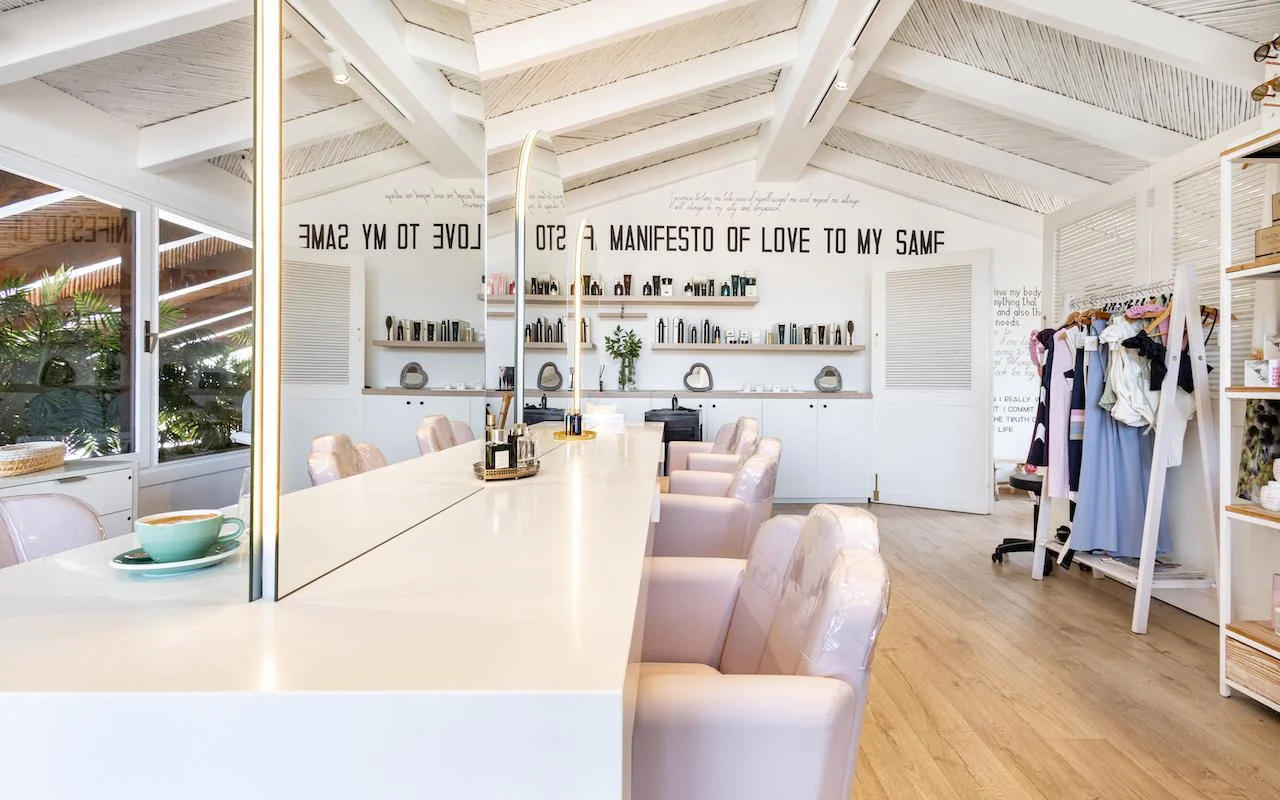 El luminoso interior de Milk & Roses en Puente Romano, Marbella, con un mostrador de peinado blanco, sillas rosa pálido, techo con vigas y estanterías de productos a lo largo de la pared.