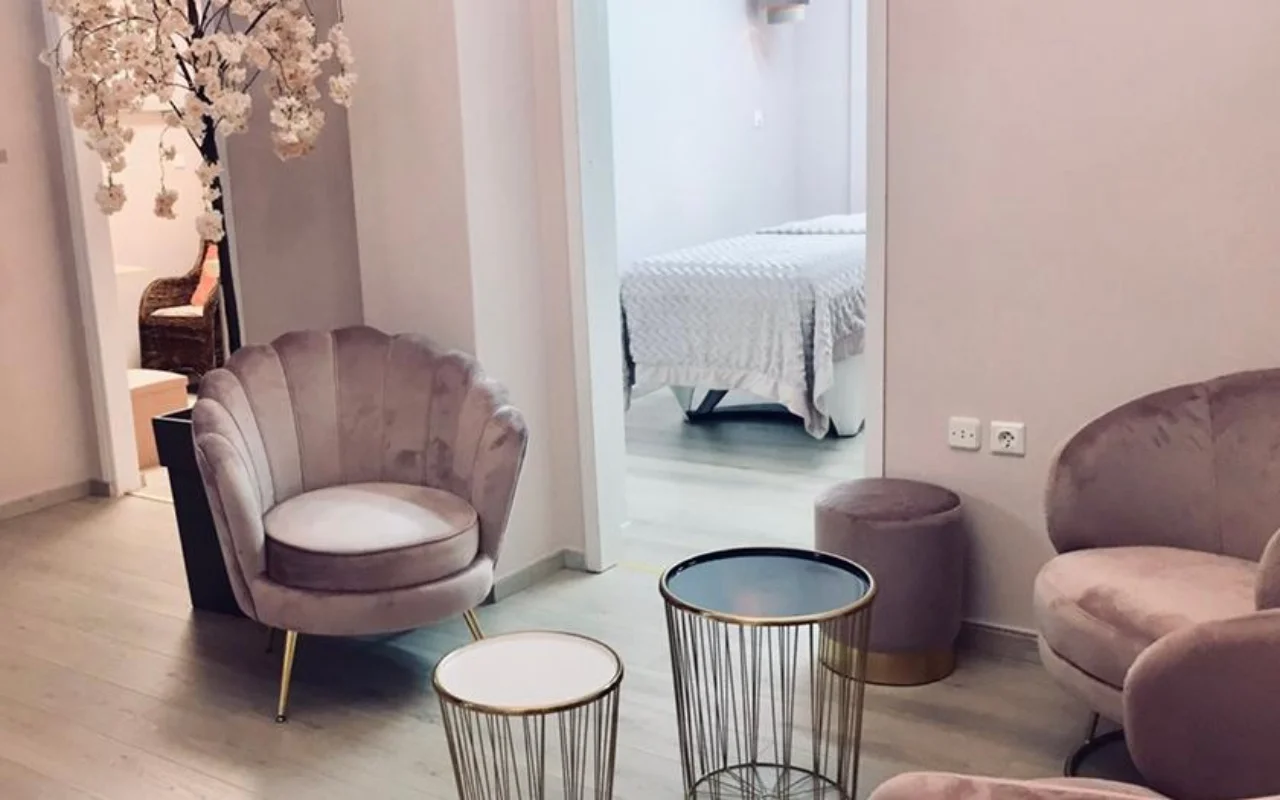 La zona de espera en rosa empolvado de Nomikou Antonia Aesthetic Lab en Syros, con una butaca de terciopelo de respaldo festoneado, finas mesas auxiliares doradas, una rama de cerezo en flor y una cabina de tratamiento al otro lado de la puerta.