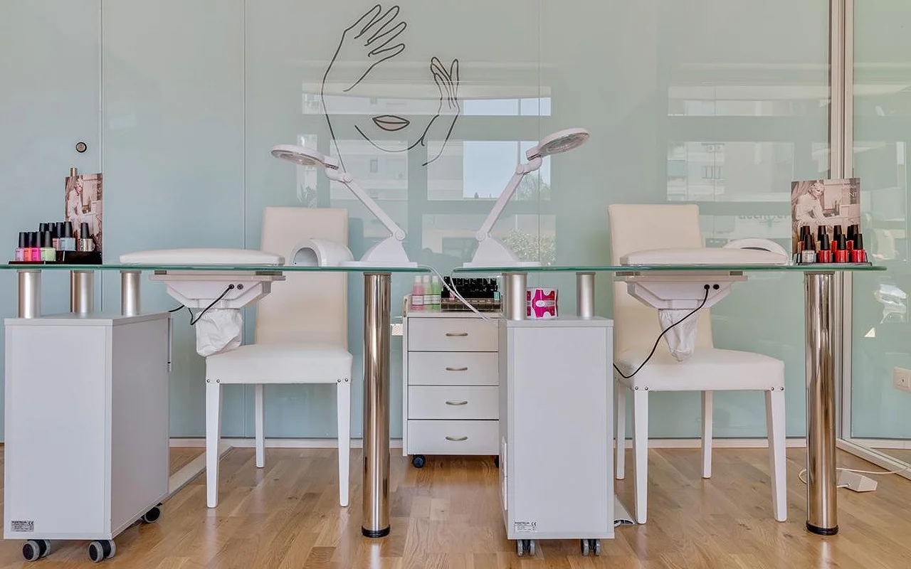 Un estudio de uñas en Perfetta Urban Beauty, con dos mesas de manicura de sobre de cristal y sillas blancas, lámparas de trabajo, expositores de esmaltes de colores y una pared de cristal verde menta con el dibujo a línea de una cara y unas manos.