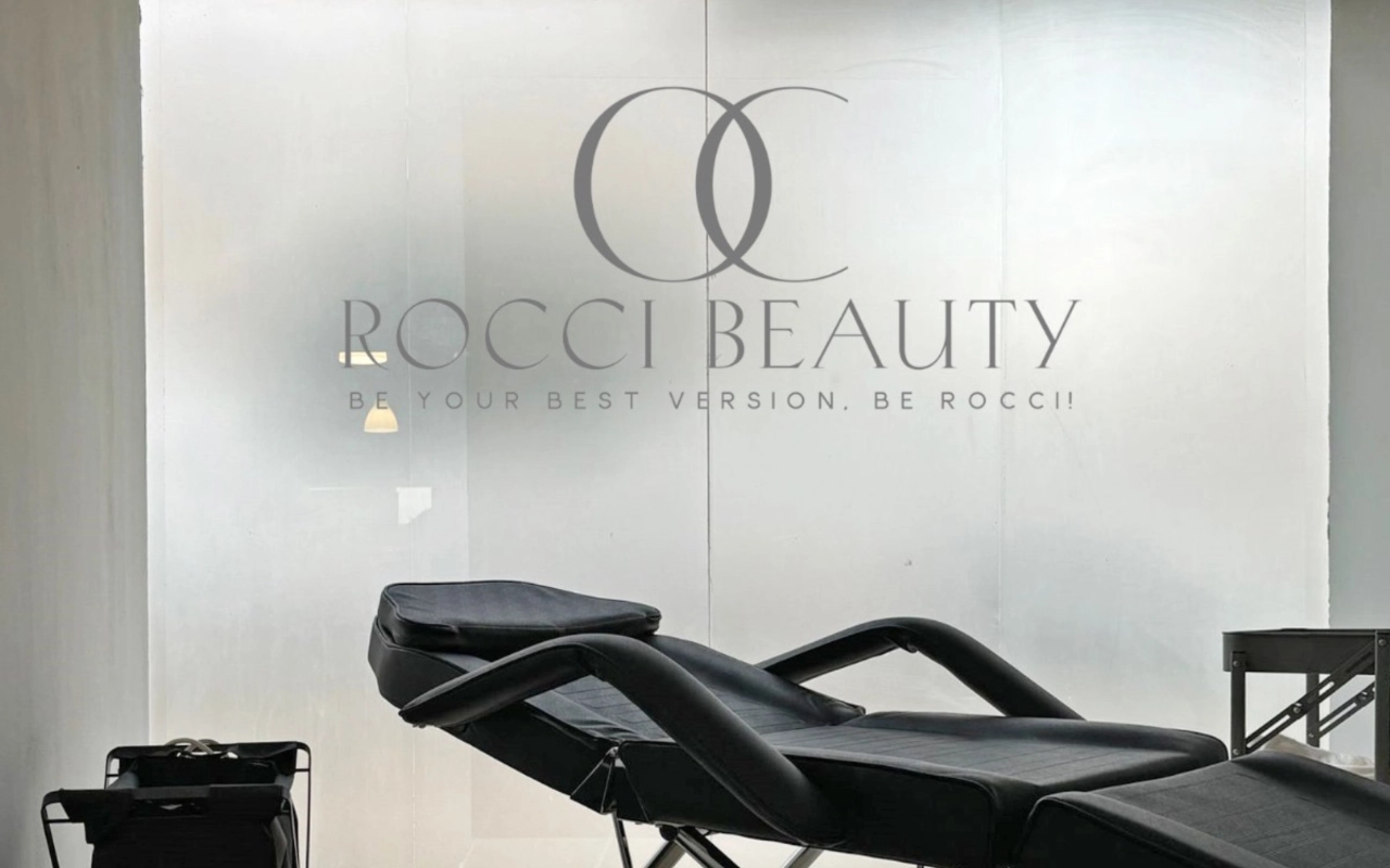 Una sala de tratamientos con una camilla reclinable negra y un pequeño carrito de equipo, ante una mampara de vidrio esmerilado que luce el logo y el lema de Rocci Beauty.