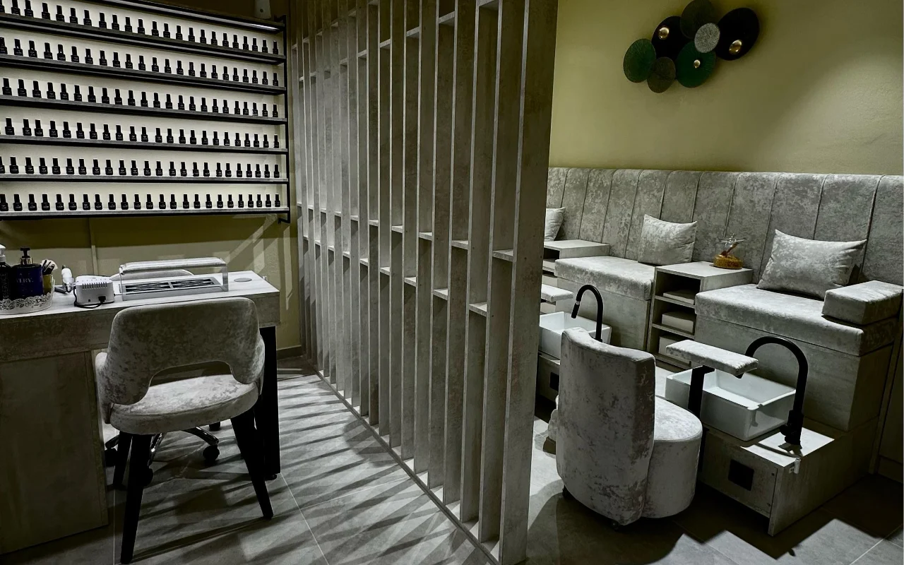 Una cabina de tratamiento en Sisu Nail Club and Academy en Perissa, Santorini, con una pared entera de esmaltes, sillones de terciopelo gris para manicura y pedicura y un separador de hormigón calado.