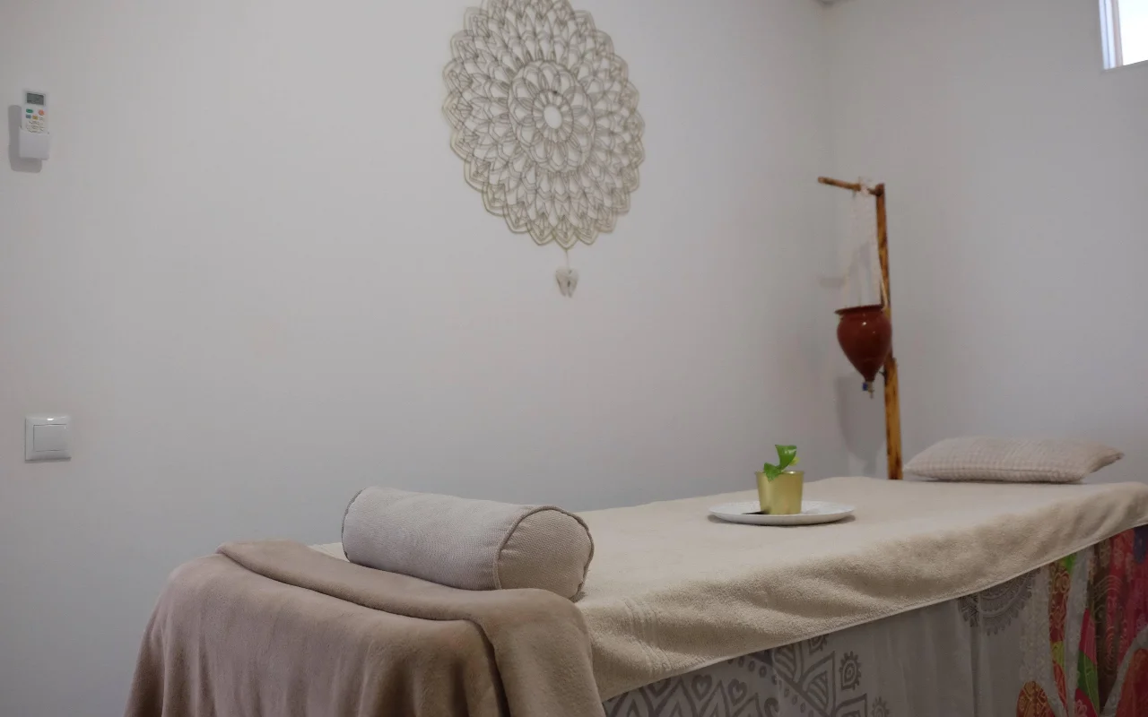 Una sala de masaje tranquila en tonos neutros y suaves, con una camilla cubierta con una toalla y un rodillo, un gran mandala de ganchillo colgado en la pared y un soporte de madera que sostiene una maceta de terracota suspendida.