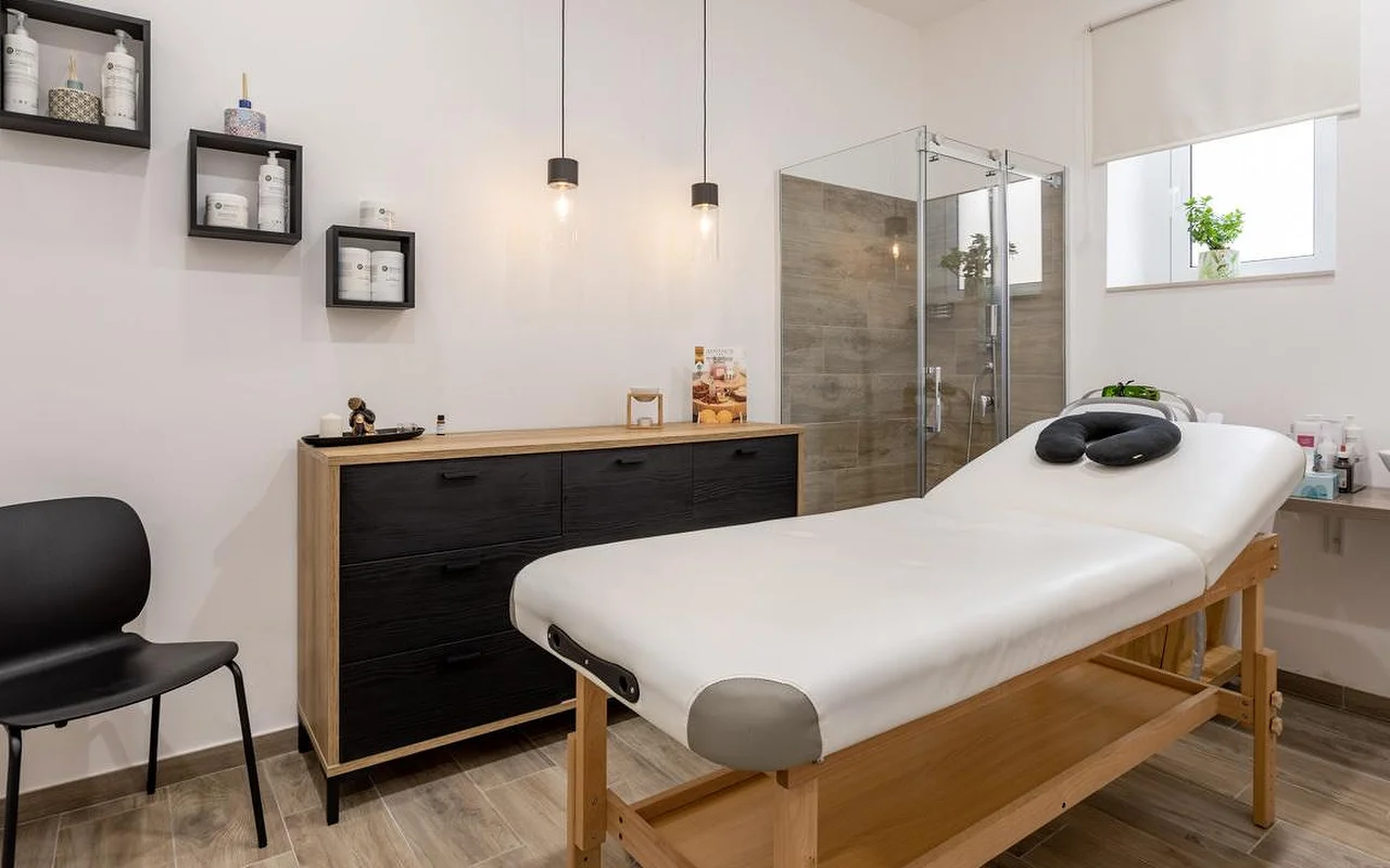 Una sala de masajes en Thea Estetica, con una camilla de masaje de madera rematada con un reposacabezas negro, una cómoda en negro y madera, dos lámparas colgantes, estanterías cubo con productos y una mampara de ducha de cristal con azulejos efecto madera.