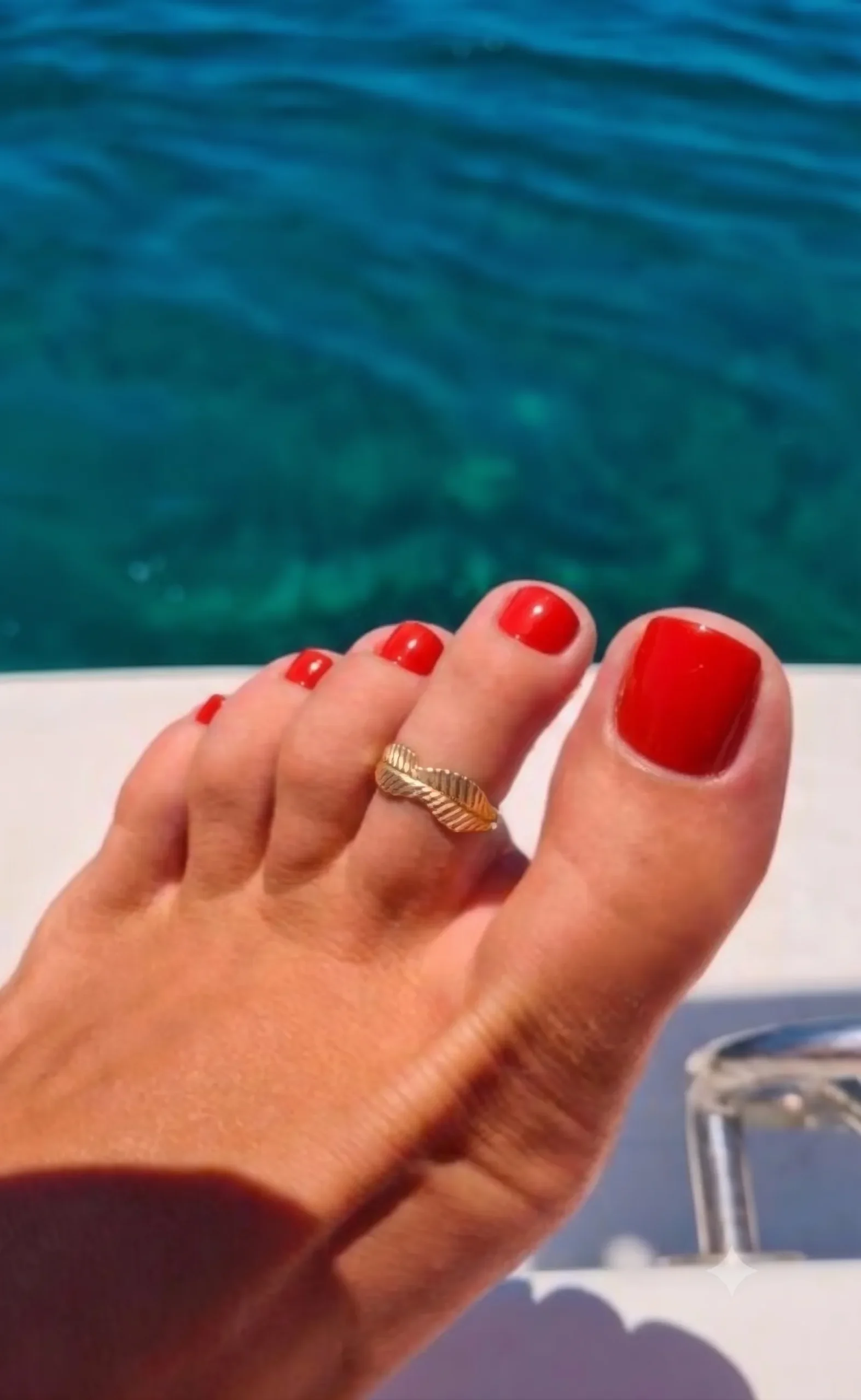 Pedicura en rojo tomate brillante sobre un pie bronceado con un anillo de pie dorado, mar turquesa de fondo
