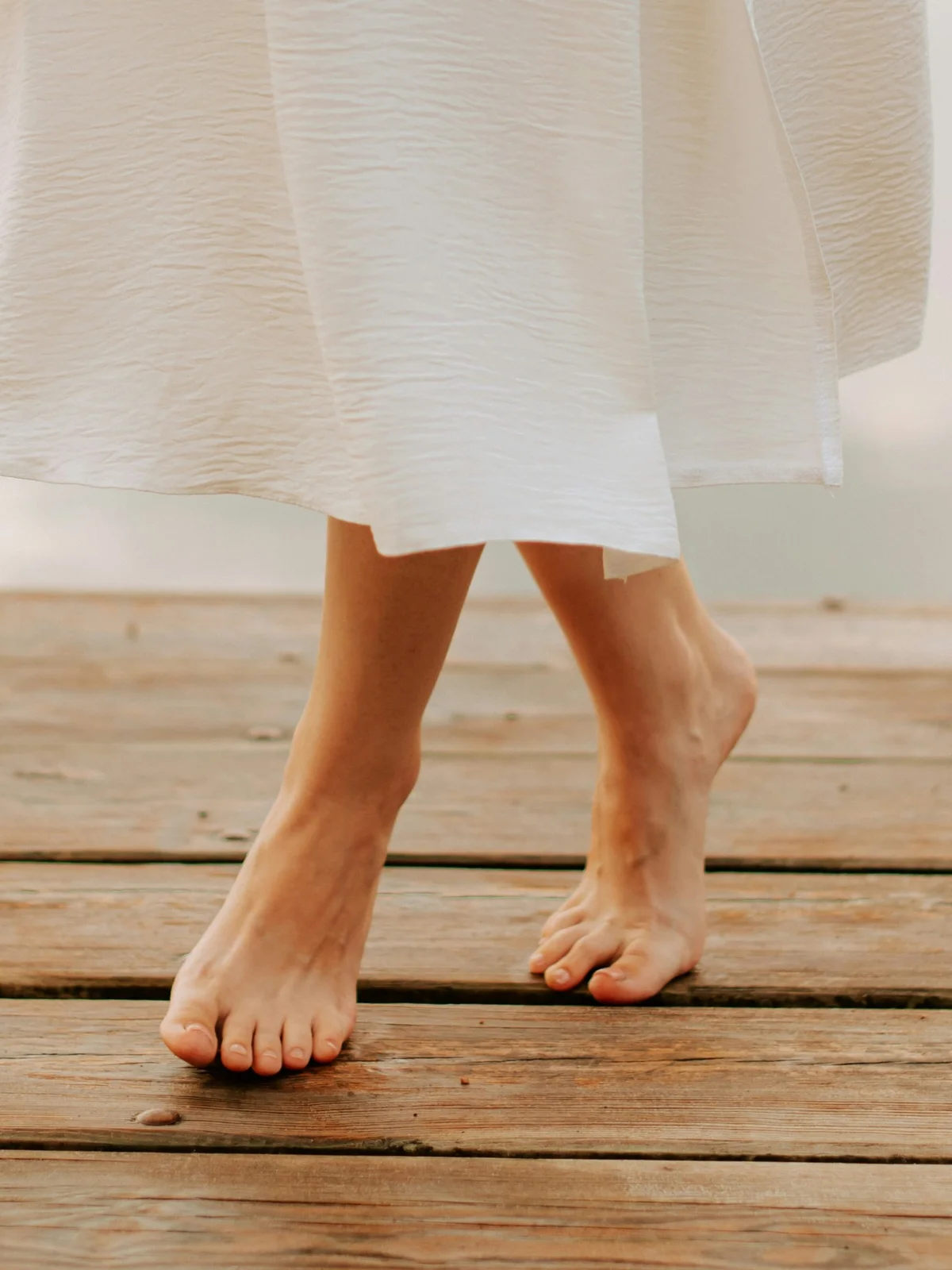 Primer plano de las piernas de una mujer con los pies descalzos y sanos y una falda blanca vaporosa, dando un paso adelante sobre una pasarela de madera junto a un lago