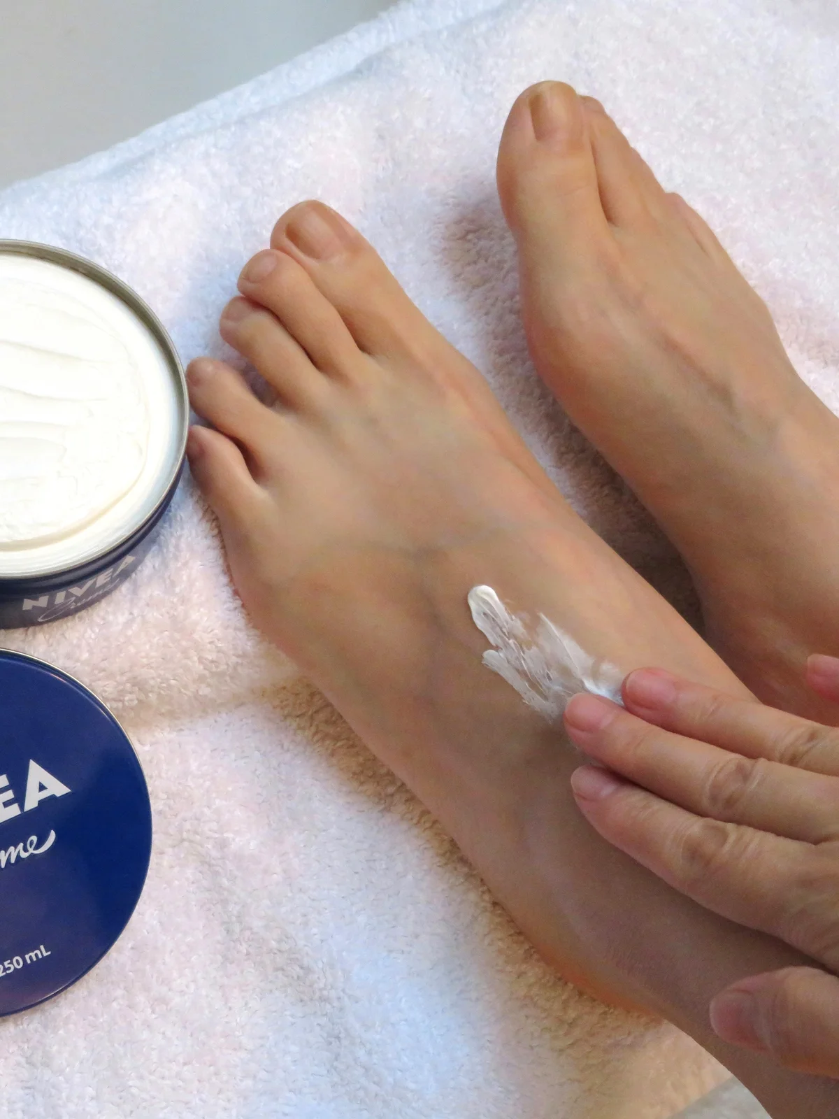 Primer plano desde arriba de los pies de una mujer apoyados sobre una sábana blanca, con un bote de crema abierto al lado, mientras se aplica la crema