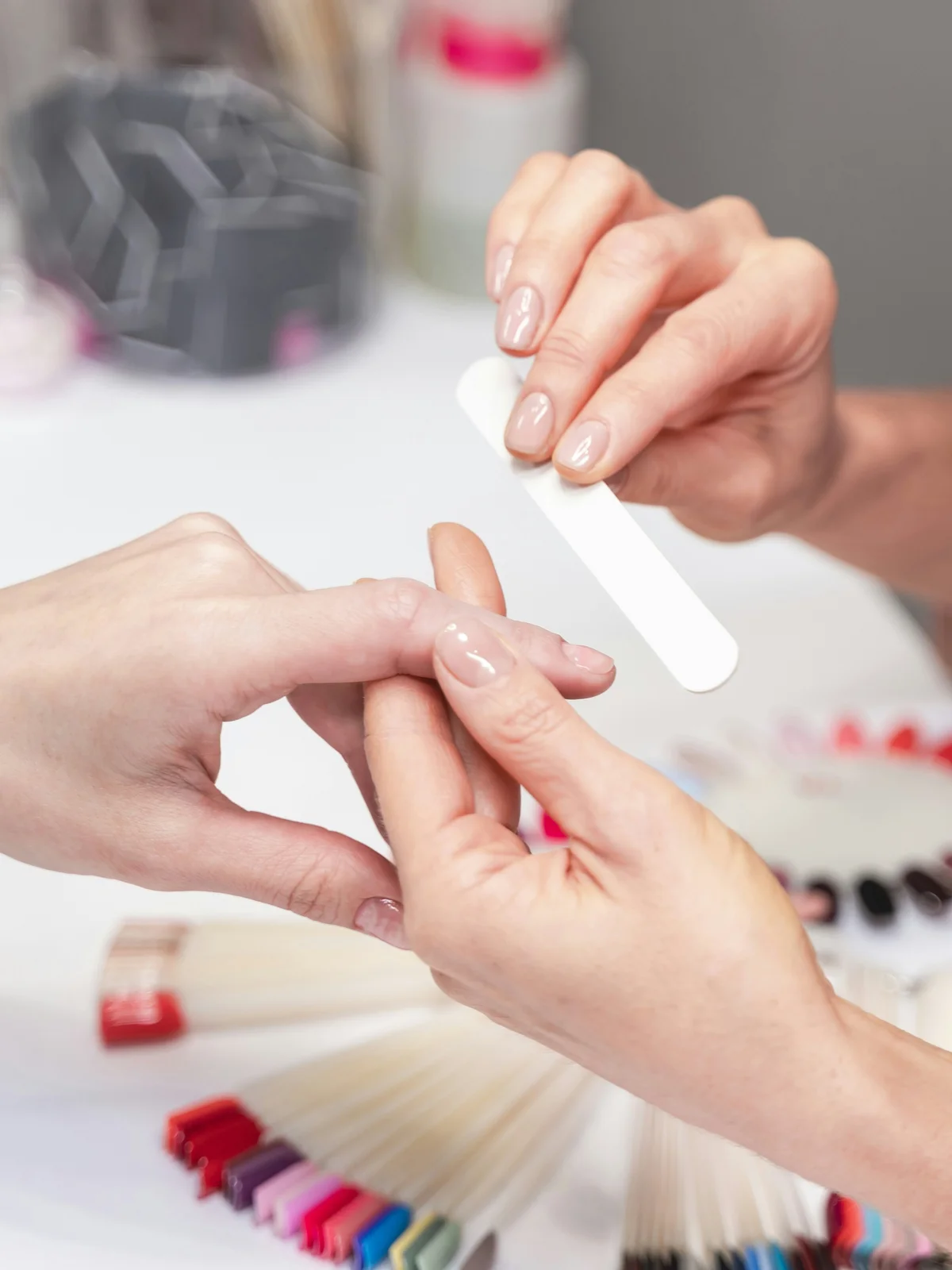 Primer plano de una manicurista limando con suavidad las uñas de una clienta en el centro, con productos y utensilios de manicura al fondo