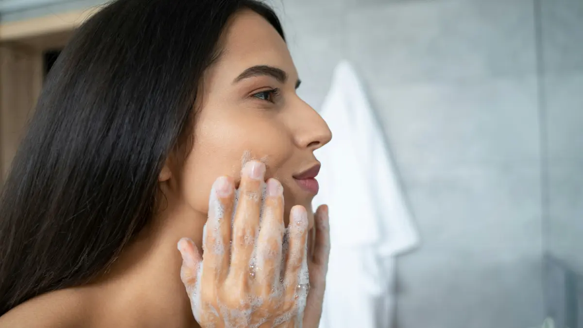 Vista de perfil de una mujer que se masajea un limpiador en la cara, inclinada sobre el lavabo del baño