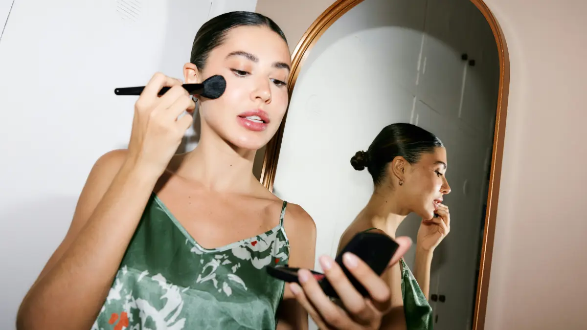 Una mujer elegante y lista para salir se mira en un espejo de bolsillo mientras se aplica maquillaje con una brocha