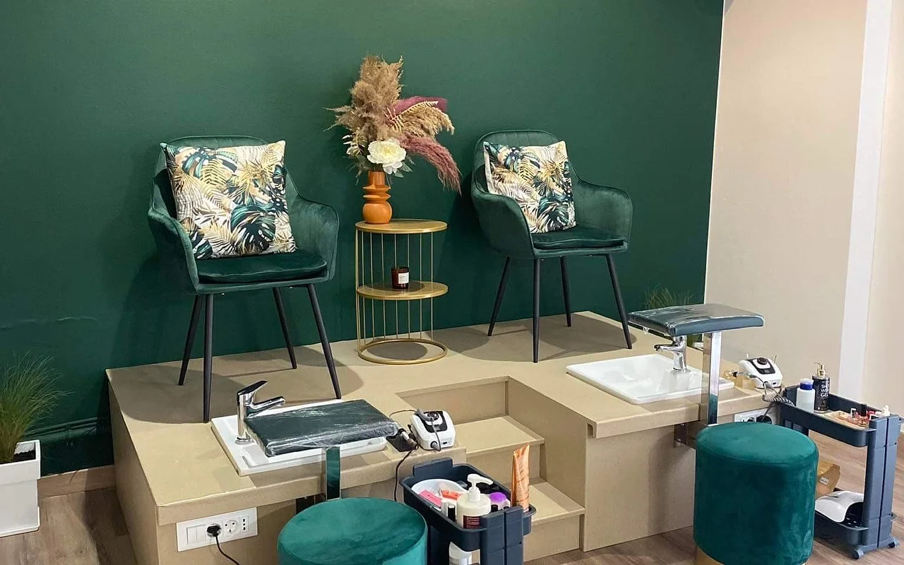 Un rincón de pedicura con una pared verde intenso, dos sillones tipo cubo en terciopelo verde con cojines de estampado tropical elevados sobre una plataforma beige encima de barreños de pedicura, una mesita auxiliar dorada con pampas en un jarrón naranja, y taburetes a juego en terciopelo verde.
