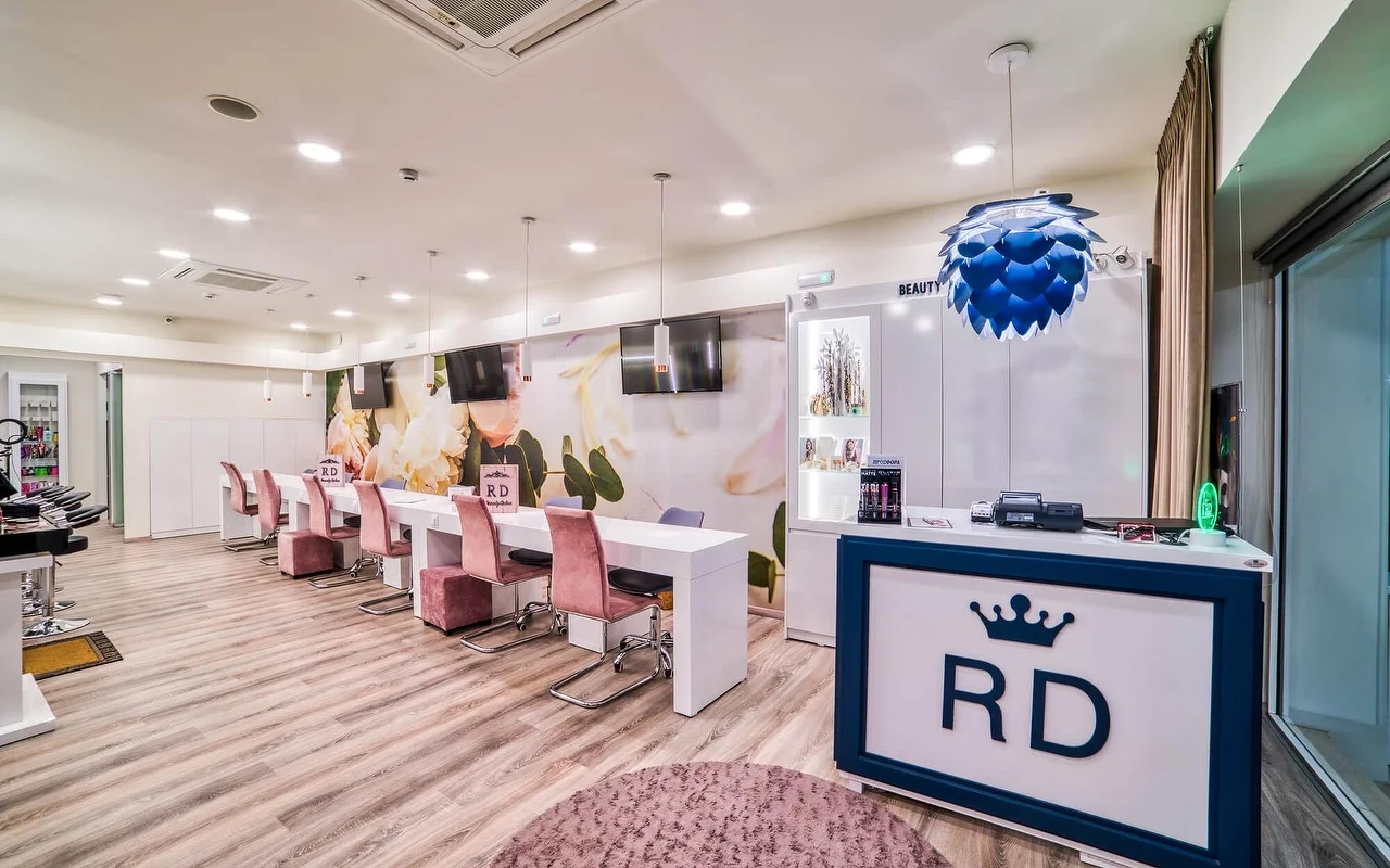 Un salón blanco y luminoso con una larga hilera de mesas de manicura y sillas rosa palo, un delicado mural floral, suelo de madera clara, y un mostrador de recepción marcado con un logo de corona 'RD'.