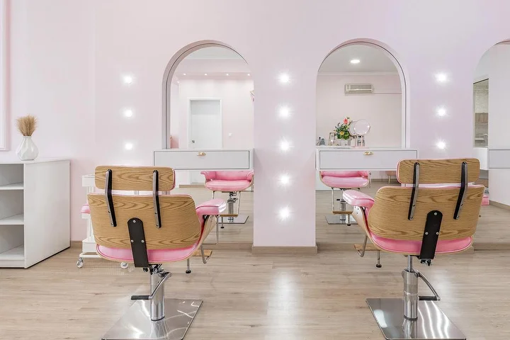 Peluquería luminosa en rosa suave, con sillones de peluquería en madera y tapicería rosa frente a dos hornacinas de arco iluminadas con focos, mobiliario blanco y suelo con efecto madera clara.