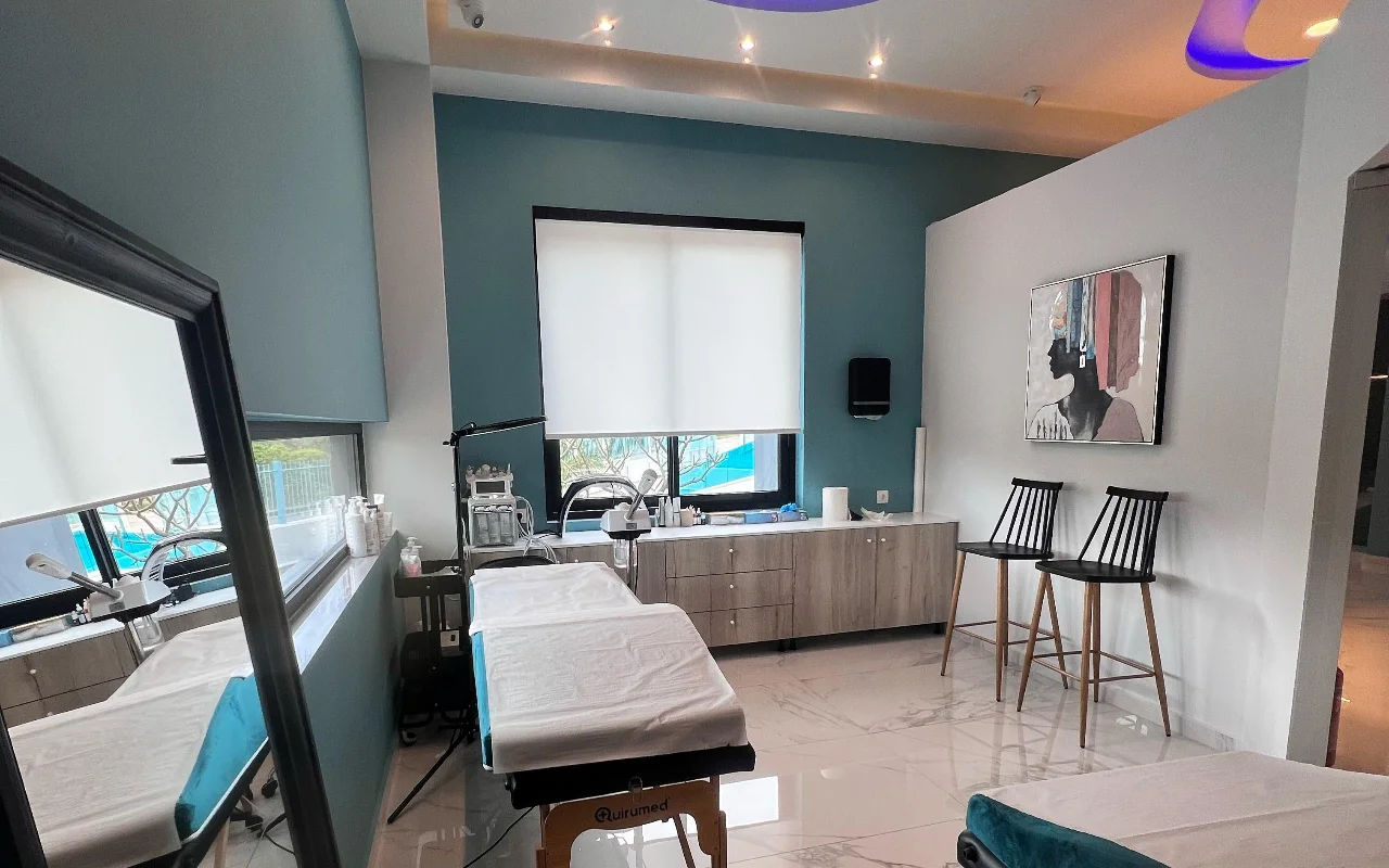 Una sala de tratamiento facial con paredes verde azulado, una camilla de tratamiento blanca, aparatos de skincare, un gran espejo, dos taburetes negros de listones y un retrato enmarcado, iluminada por una ventana con estor enrollable blanco.