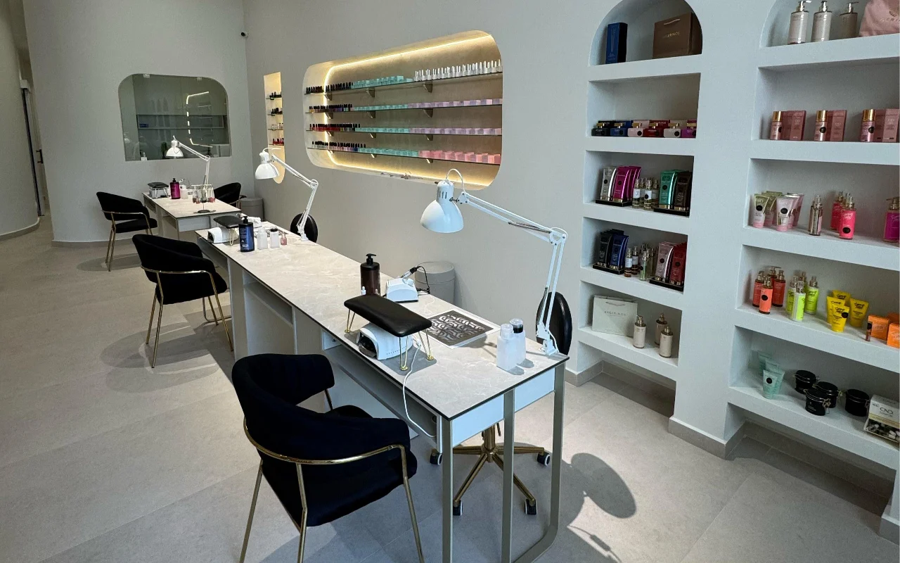 Un estudio de uñas blanco con mesas de manicura de tapa de mármol y lámparas articuladas blancas, sillas de terciopelo negro sobre patas doradas, y hornacinas arqueadas en la pared que exhiben productos de skincare y un expositor retroiluminado de esmaltes.