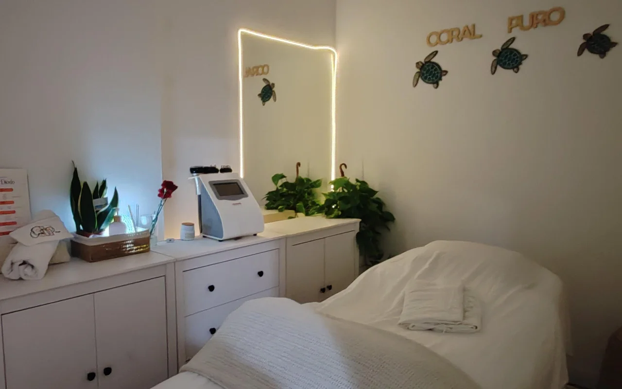 Una pequeña y tranquila sala de tratamientos con armarios blancos y una camilla vestida con toallas blancas. Un espejo iluminado y una máquina de tratamiento están sobre el mostrador entre plantas verdes, bajo un letrero dorado que reza «Coral Puro» y arte mural decorativo en forma de tortuga.
