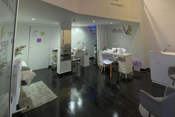Interior de un centro de belleza: suelo de baldosas oscuras y pulidas, mesa de manicura blanca y áreas de tratamiento con luz tenue, el logo Mulher en la pared del fondo y una camilla facial visible tras una pared de cristal.